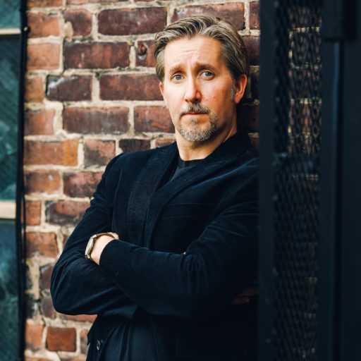 Josh Boldt leaning against brick wall wearing black blazer and t-shirt